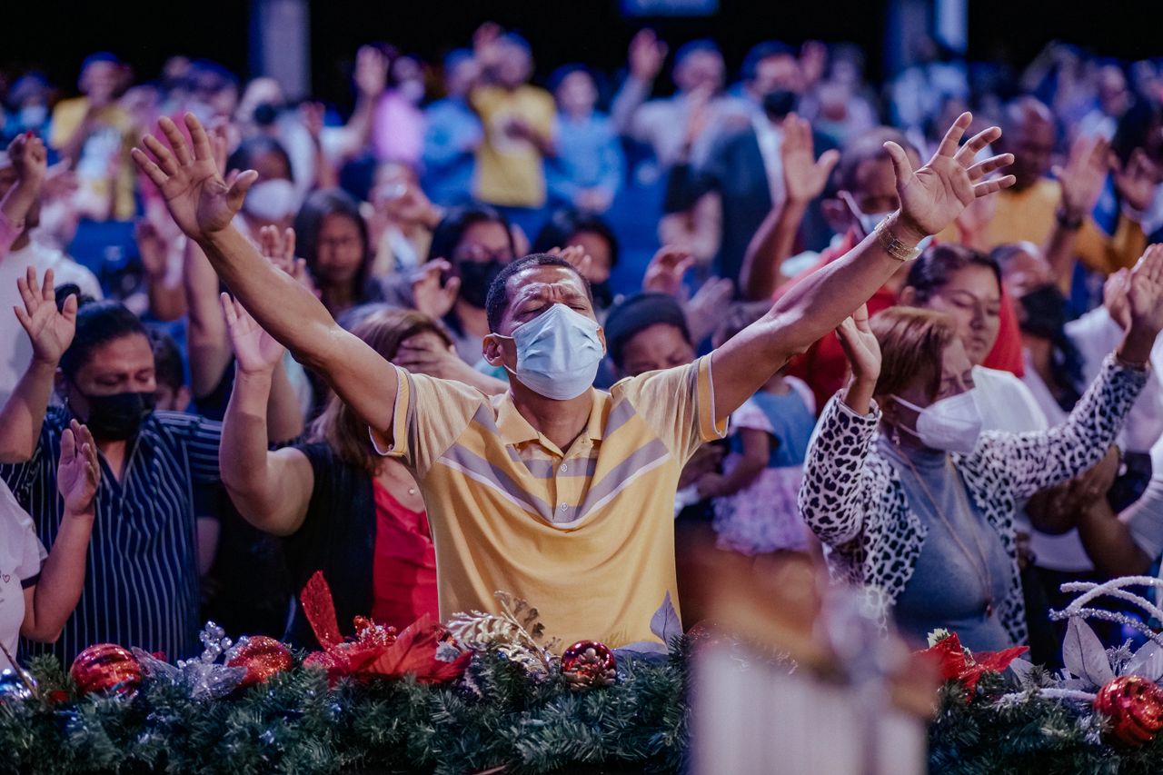Diverse group of people raise their hands during a worship gathering indoors while masked.